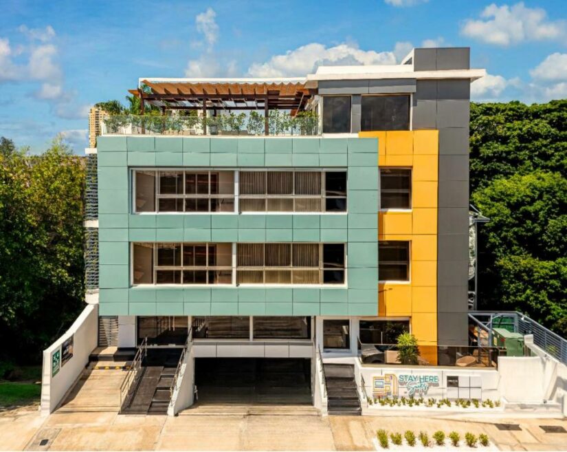 Edifício moderno de três andares com fachada colorida, terraço na cobertura e garagem, cercado por vegetação.