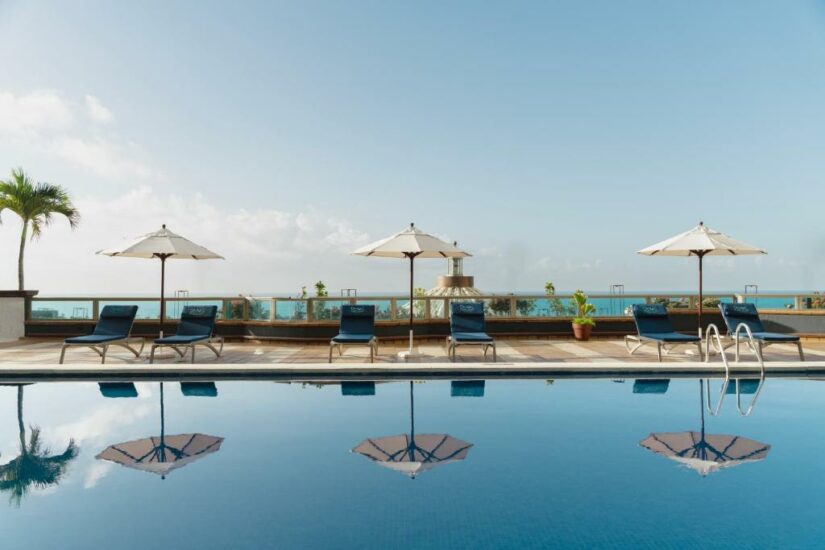 Uma fileira de espreguiçadeiras e guarda-sóis em uma piscina azul clara com vista para o oceano sob um céu ensolarado.