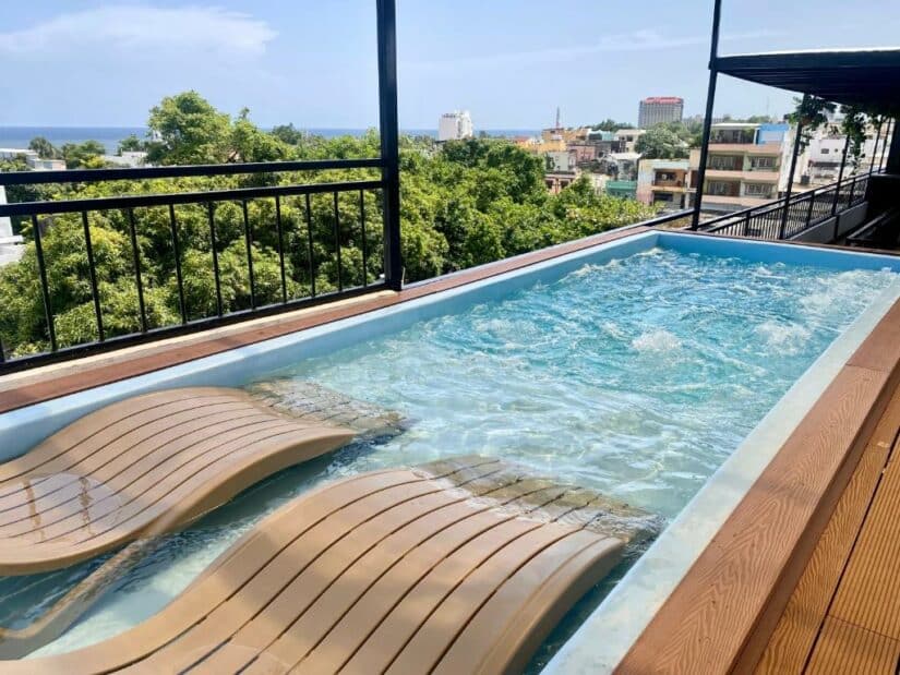 Piscina na cobertura onde ficar em Santo Domingo com duas espreguiçadeiras de madeira, prédios da cidade e vegetação ao fundo sob um céu azul.
