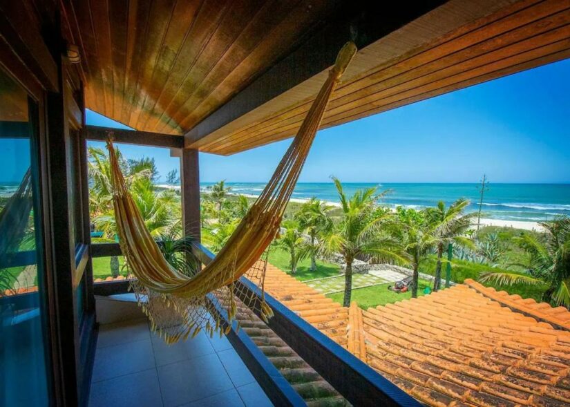 Uma rede em uma varanda onde ficar em Saquarema com vista para palmeiras, telhados e uma praia ensolarada com céu azul e oceano.