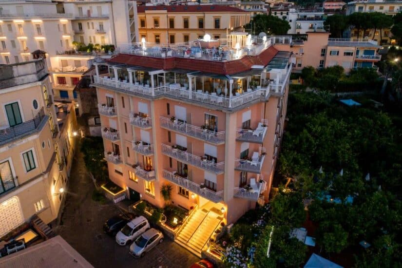 Um hotel onde ficar em Sorrento cor de pêssego com varandas e assentos na cobertura, iluminado ao anoitecer, com carros estacionados na frente.