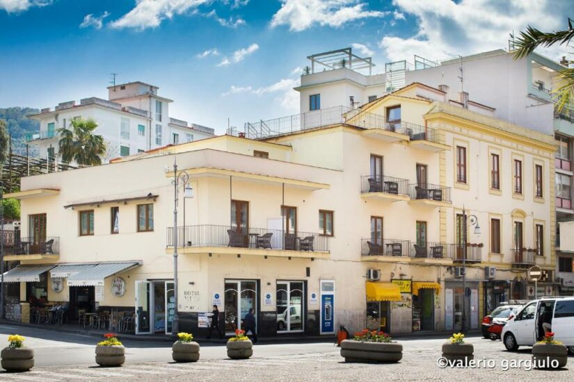 Dia ensolarado em uma cidade europeia com um café de esquina, um hotel e uma loja de toldos amarelos, cercada por palmeiras.