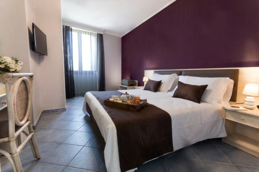 Quarto de hotel aconchegante com cama de casal, parede com detalhes em roxo, bandeja com café da manhã e uma janela com cortinas.