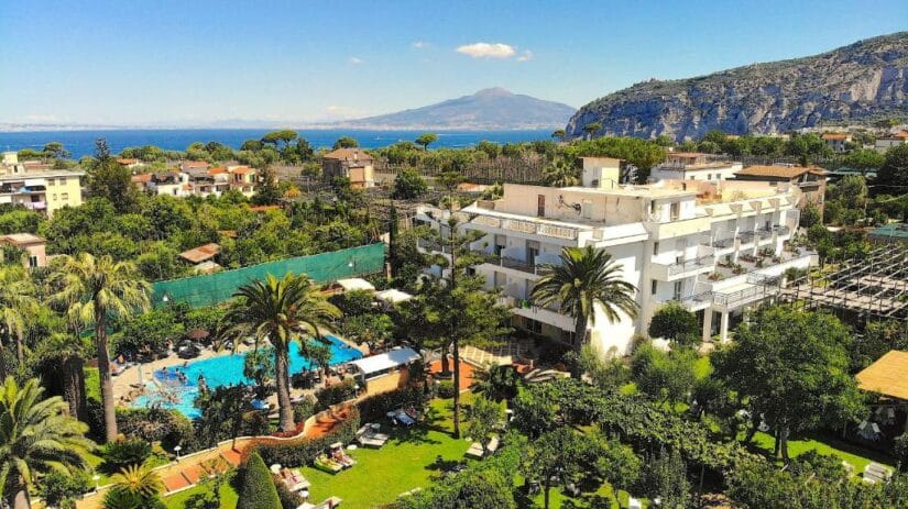 Vista aérea de um hotel onde ficar em Sorrento com piscina, jardins exuberantes, montanhas e mar ao fundo.