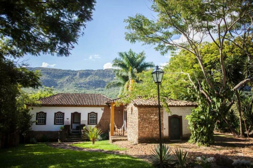 Casa em estilo colonial com paredes brancas e telhado de telha, cercada por árvores, com montanhas ao fundo.
