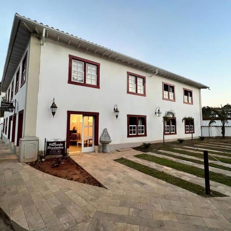 Edifício branco de dois andares onde ficar em Tiradentes com acabamento vermelho, porta aberta, passarela de pedra e pequenas plantas na frente.
