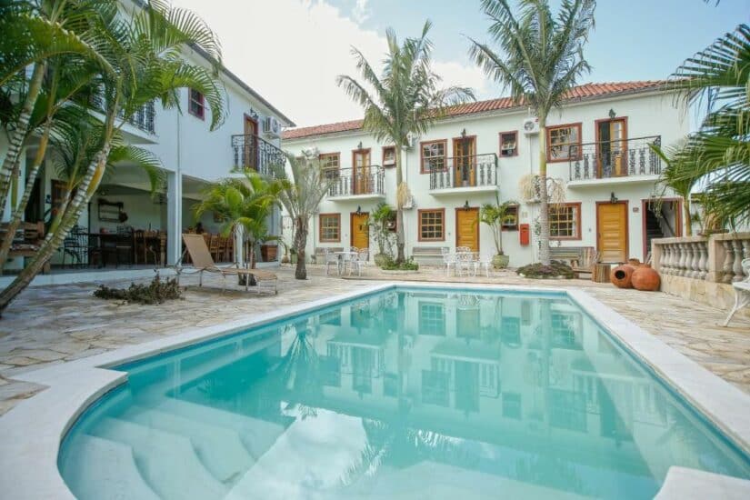 Pátio com palmeiras, piscina, espreguiçadeiras e prédio branco de dois andares com varandas e telhado de telhas vermelhas.
