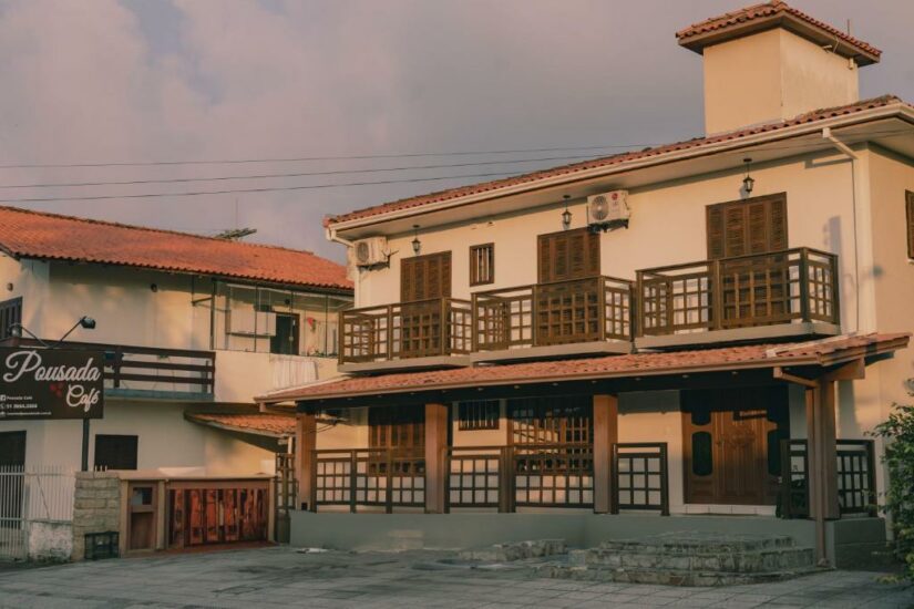 Prédio de dois andares onde ficar em Torres com varandas de madeira e uma placa indicando Pousada Café sob um céu quente de fim de tarde.