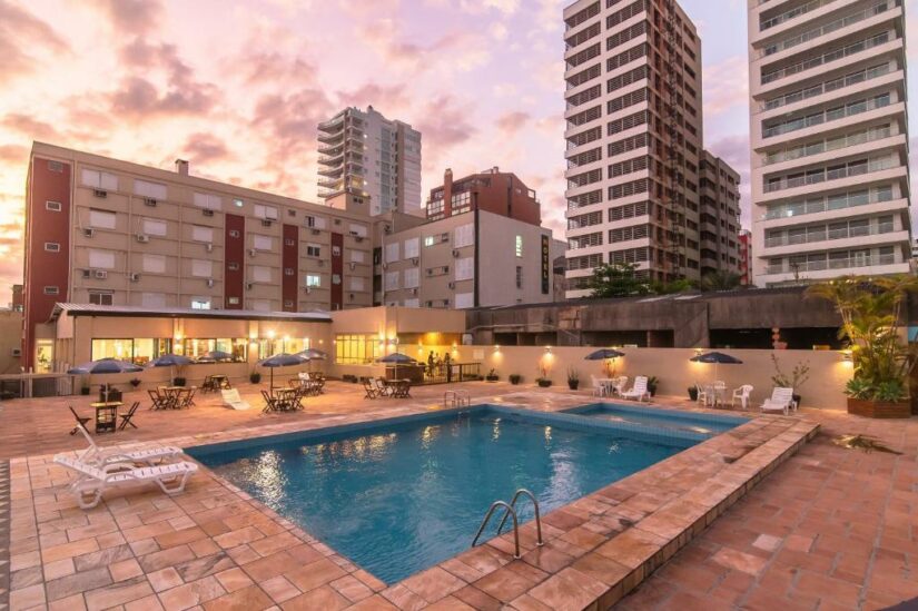 Uma piscina cercada por espreguiçadeiras e mesas, com prédios altos ao fundo no pôr do sol.