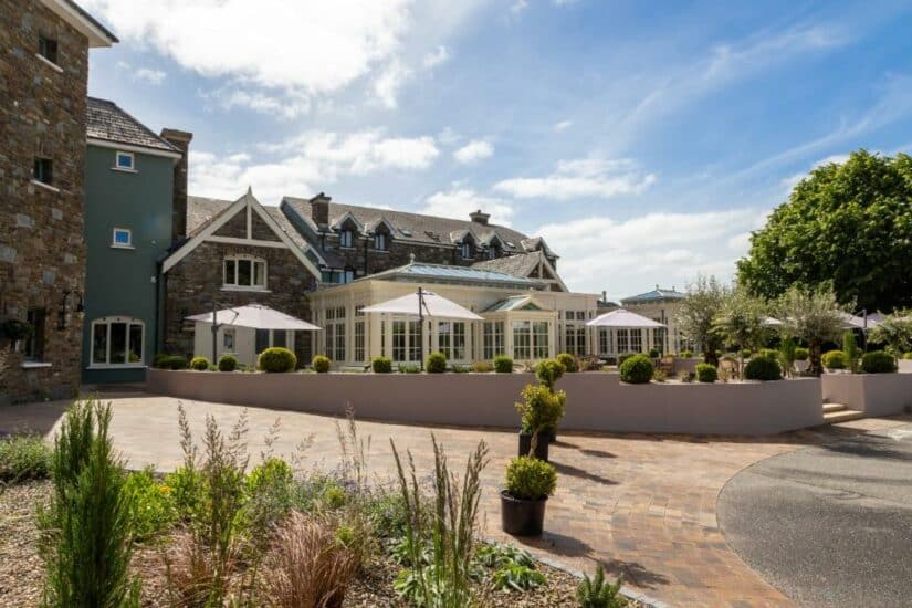Elegante hotel de pedra com grandes janelas onde ficar na Irlanda, pátio externo e jardim sob um céu azul com nuvens dispersas.