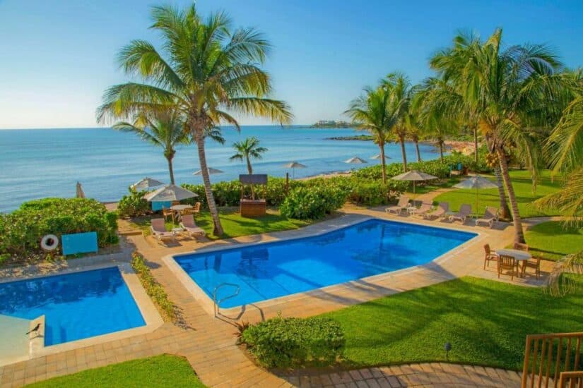 Área da piscina do resort com palmeiras, espreguiçadeiras e vista para o mar sob um céu azul claro.