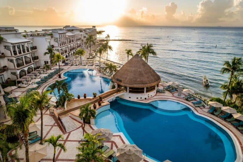 Vista aérea de um resort à beira-mar onde ficar na Riviera Maya com piscinas, palmeiras e oceano ao nascer do sol.