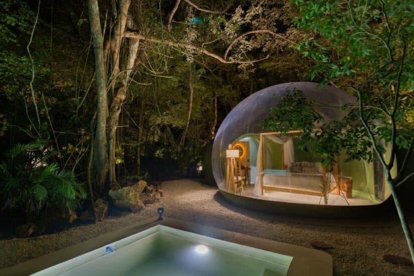 Uma tenda de domo transparente com uma cama dentro fica em uma floresta onde ficar na Riviera Maya, ao lado de uma pequena piscina iluminada à noite.