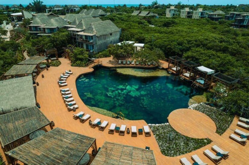 Vista aérea de um resort com uma piscina em estilo de lagoa, espreguiçadeiras e vilas com telhado de palha cercadas por vegetação.