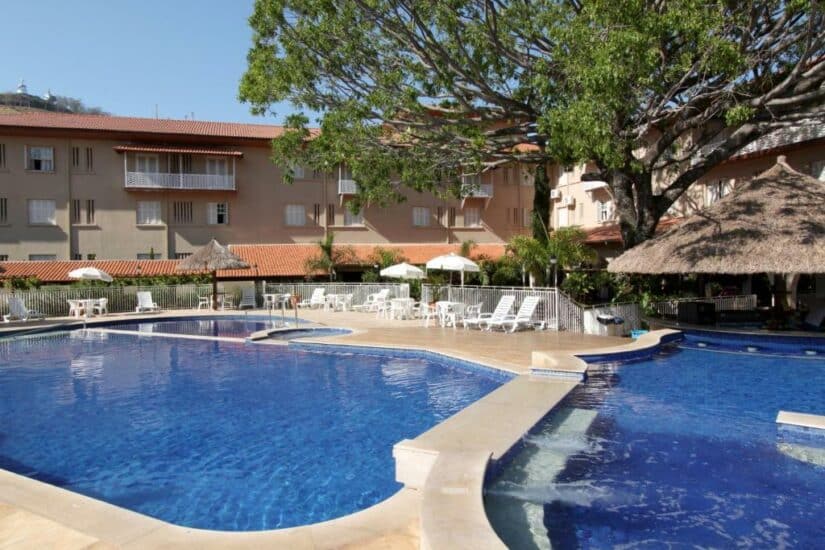 Uma piscina de hotel Onde ficar em Águas de Lindoia com espreguiçadeiras, guarda-sóis e uma grande árvore, ao lado de um prédio bronzeado.