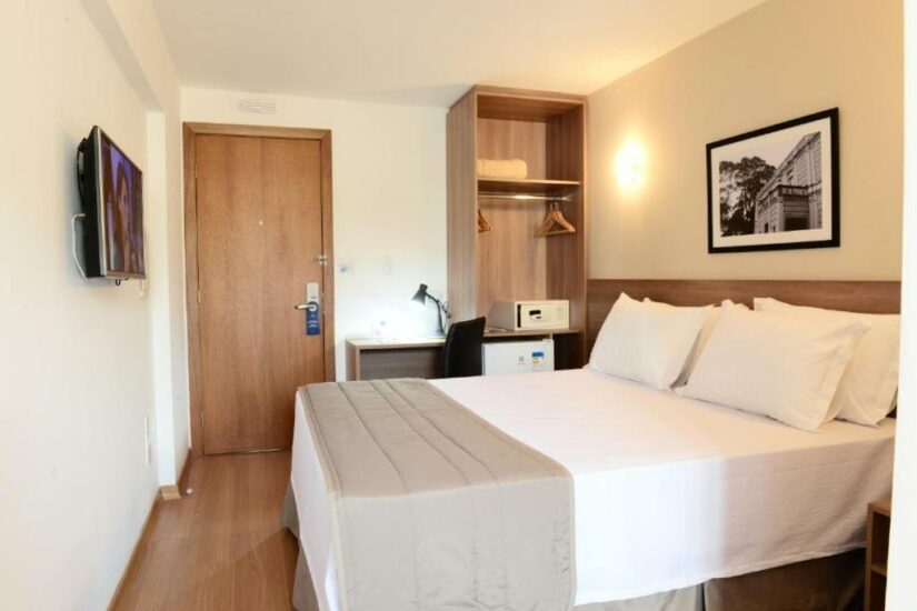 hotéis no centro de juiz de fora mg Quarto de hotel moderno Onde ficar em Juiz de Fora com uma cama de casal bem arrumada, escrivaninha, armário, TV e um quadro emoldurado na parede.