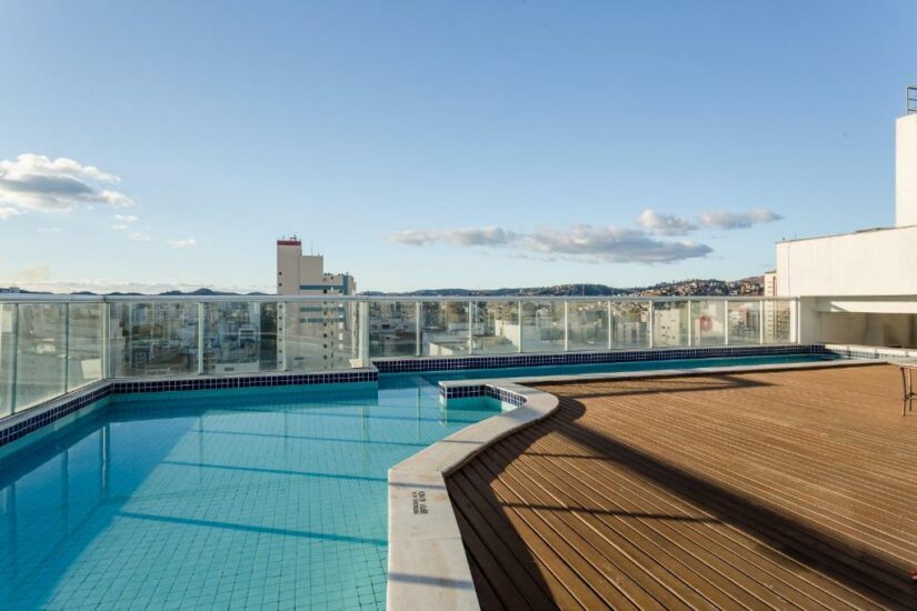 Onde ficar em Juiz de Fora MG Piscina na cobertura com deck de madeira, prédios da cidade e colinas ao fundo sob um céu claro.