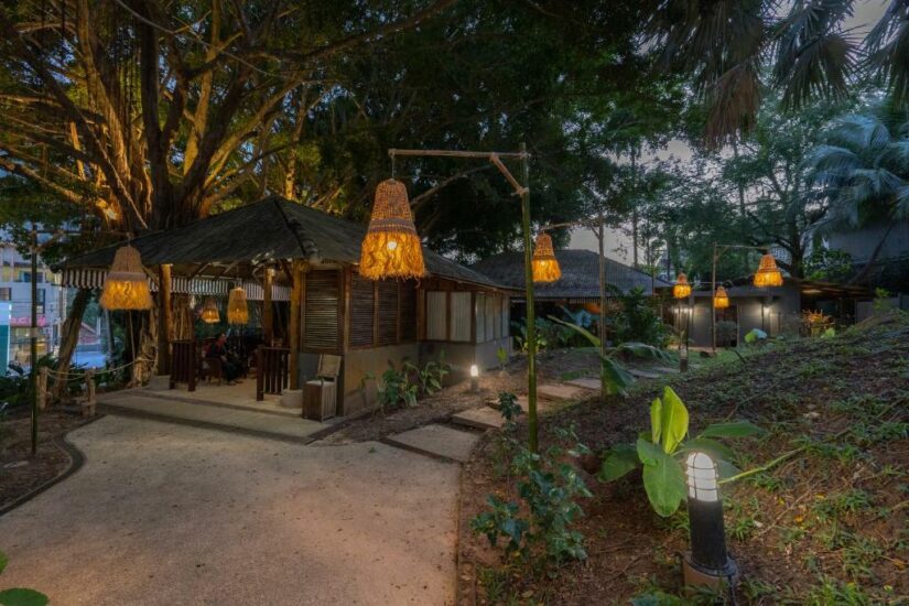 Caminho ao ar livre com lanternas penduradas, vegetação exuberante e cabanas de madeira aconchegantes sob grandes árvores ao anoitecer onde ficar em Krabi.