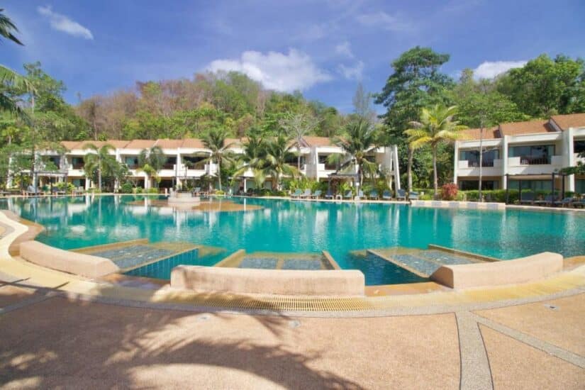 Piscina do resort com águas claras, cercada por palmeiras e prédios brancos sob um céu azul.