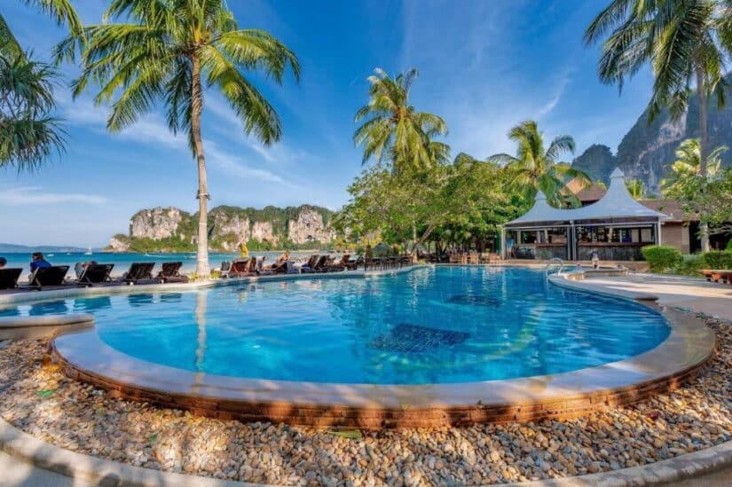 Uma piscina redonda de resort com palmeiras, espreguiçadeiras e uma praia com falésias ao fundo sob um céu azul.