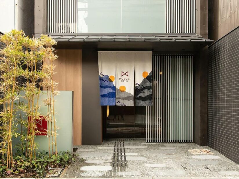 Entrada de um edifício moderno onde ficar em Kyoto com o banner do Musubi Hotel, plantas de bambu e caminho de pedra.