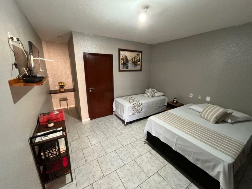 Quarto de hotel com duas camas, uma mesa pequena, TV em uma prateleira na parede e uma porta que leva a outro quarto.