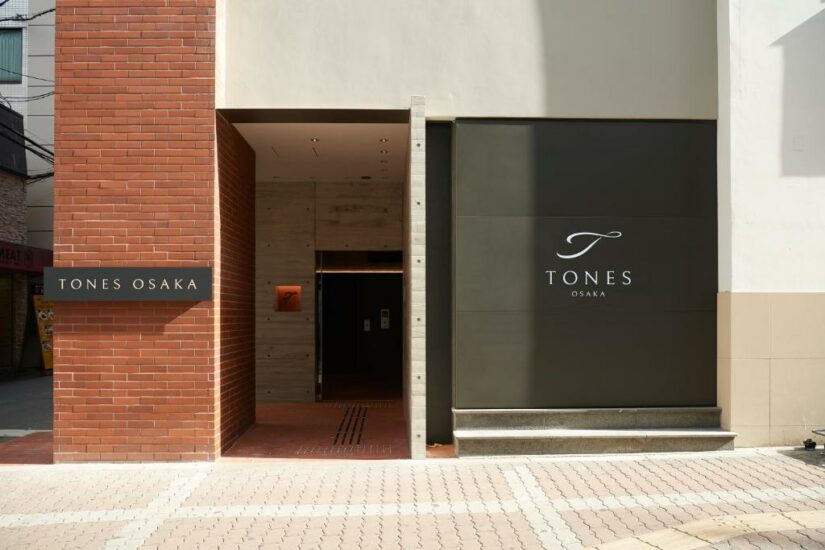 Entrada da TONES com paredes de tijolos e cinza, e a placa da TONES ao lado da porta.