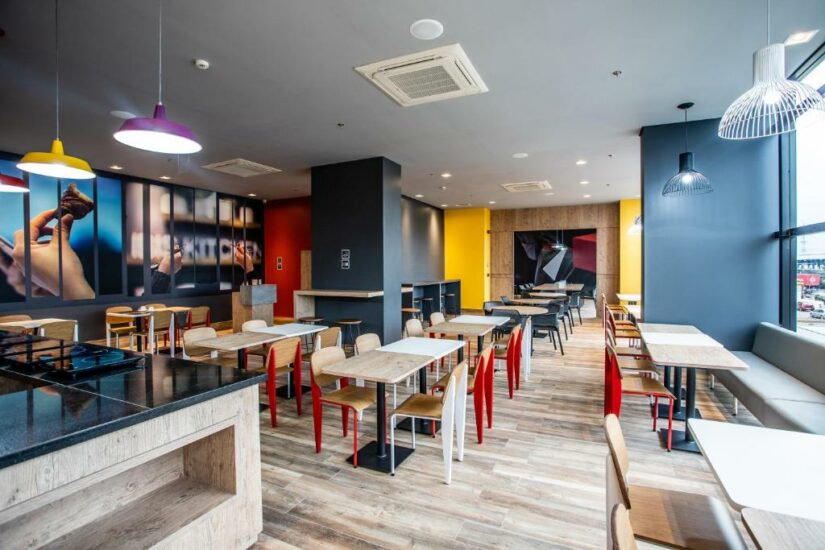 Interior de cafeteria moderna com mesas de madeira, cadeiras coloridas e iluminação decorativa, com fotos grandes na parede onde ficar em Palmas.