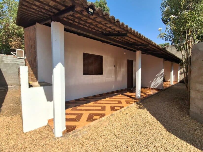 Uma pequena casa onde ficar em Palmas com paredes brancas, varanda de telhas marrons e um telhado inclinado, cercada por cascalho e árvores.