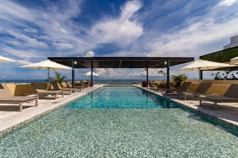Piscina infinita com espreguiçadeiras e guarda-sóis onde ficar em Playa del Carmen, com vista para o oceano sob um céu parcialmente nublado.