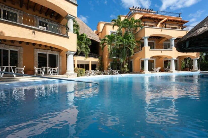 Edifício em estilo resort com varandas e palmeiras ao redor de uma piscina azul clara sob um céu ensolarado.