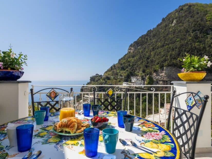 Mesa de café da manhã com pratos coloridos em uma varanda com vista para montanhas, vegetação e o mar sob um céu claro.