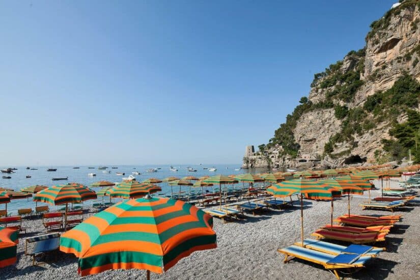 Guarda-sóis listrados coloridos e espreguiçadeiras onde ficar em Positano em uma praia de seixos com falésias e barcos no mar.