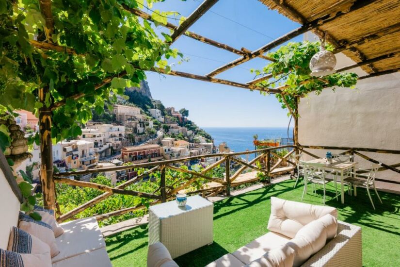 Terraço ensolarado onde ficar em Positano com videiras, móveis brancos, vista para o mar e prédios na encosta de uma cidade costeira do Mediterrâneo.