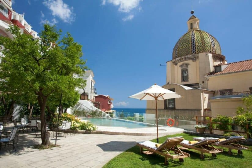 Piscina com borda infinita e espreguiçadeiras em um edifício histórico onde ficar em Positano com cúpula e vista para o mar sob um céu azul claro.
