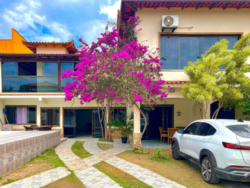 Casa moderna com janelas grandes, árvore rosa florescendo, SUV branco estacionado na entrada da garagem, céu azul acima.