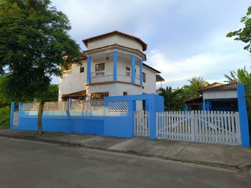 Casa branca de dois andares Onde ficar em Rio das Ostras com acabamento azul, quintal cercado e árvore na calçada sob um céu parcialmente nublado.