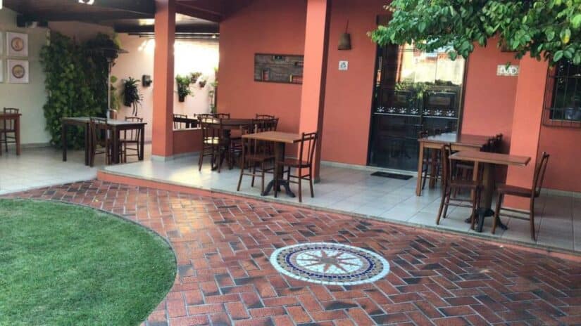 Pátio externo de uma cafeteria com mesas e cadeiras de madeira Onde ficar em Rio das Ostras, piso de tijolos e vegetação ao longo das paredes.