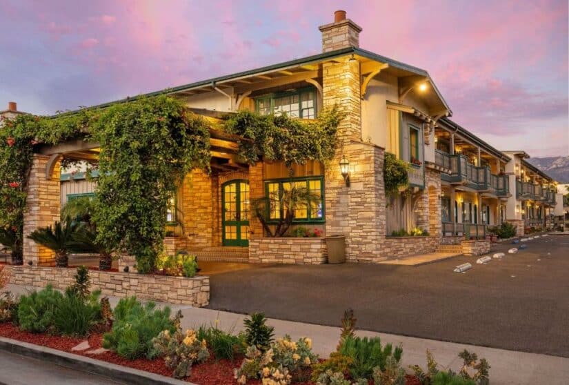 Hotel Onde ficar em Santa Barbara de dois andares com paredes de pedra, vegetação e iluminação quente ao pôr do sol sob um céu rosa e roxo.