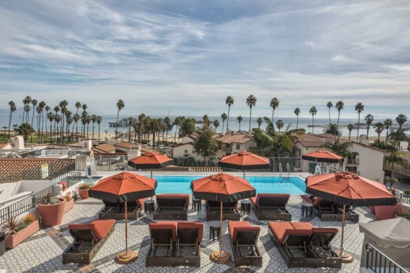 Área da piscina na cobertura com guarda-sóis vermelhos Onde ficar em Santa Barbara, espreguiçadeiras, vista para o mar e palmeiras sob um céu parcialmente nublado.