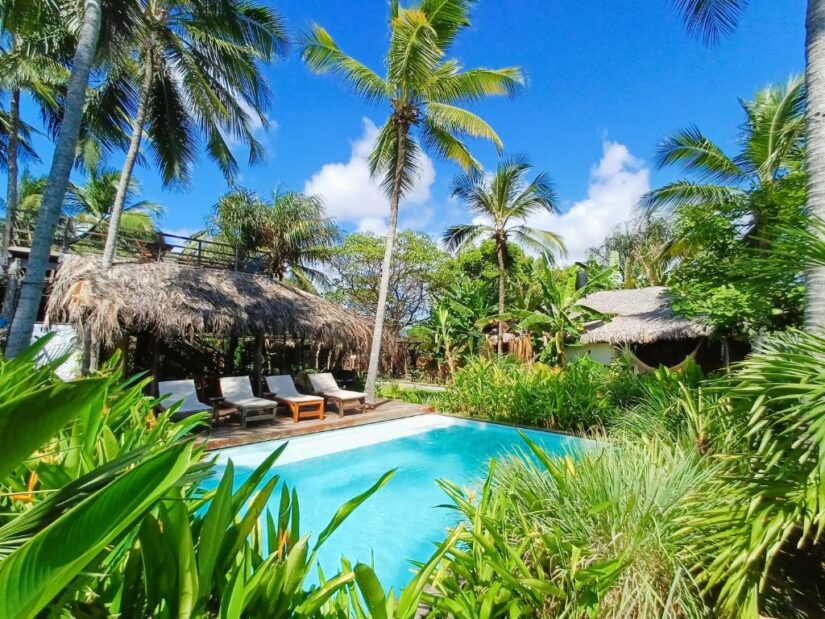 Piscina tropical cercada por uma vegetação exuberante Onde ficar em Santo Amaro do Maranhão, palmeiras, espreguiçadeiras e uma cabana com telhado de palha sob o céu azul.