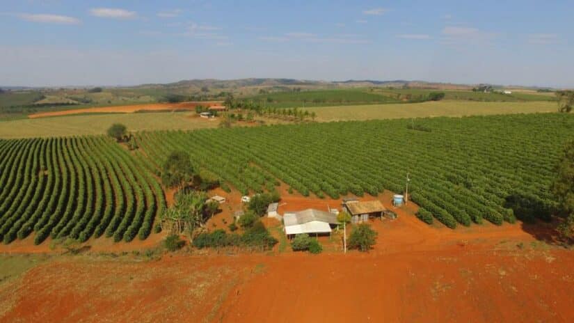 Vista aérea de uma fazenda com plantações verdes Onde ficar em São Roque de Minas, solo vermelho, pequenos edifícios e colinas ao fundo.