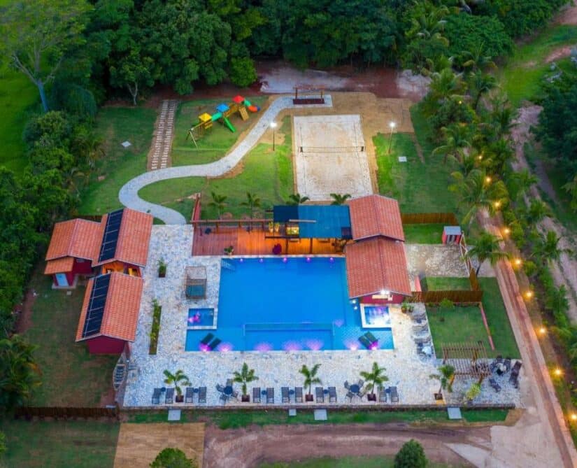 Vista aérea de uma área de piscina com espreguiçadeiras, playground, quadra de vôlei e vegetação ao redor Onde ficar em São Roque de Minas.