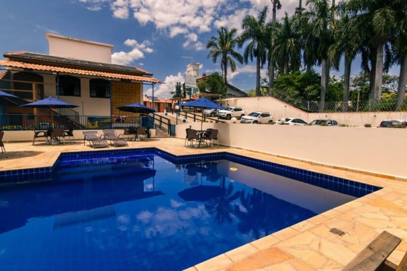 Piscina externa com água azul, espreguiçadeiras, guarda-sóis e palmeiras ao lado de um prédio onde ficar em Socorro sob um céu ensolarado.