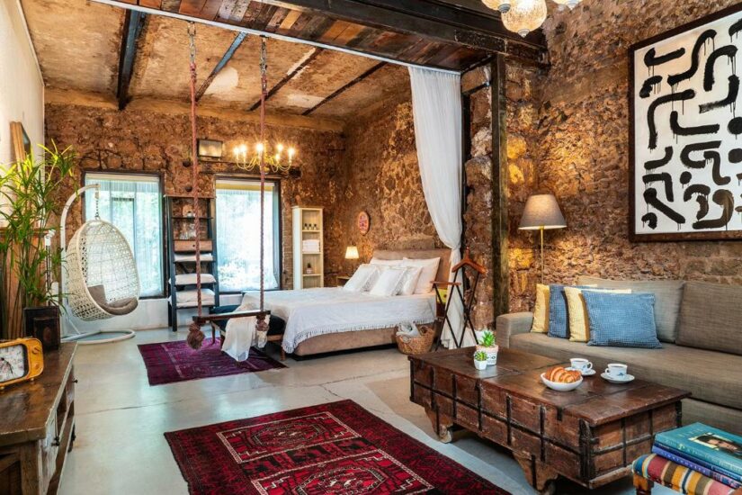Quarto rústico Onde ficar em Tel Aviv aconchegante com paredes de pedra, uma cama branca, tapetes vermelhos, um balanço e uma decoração elegante e eclética.