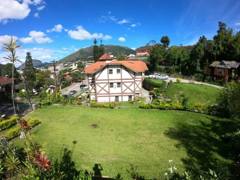 Casa em estilo enxaimel  onde ficar em Teresópolis com telhado vermelho, grande gramado verde e montanhas ao fundo sob um céu azul.