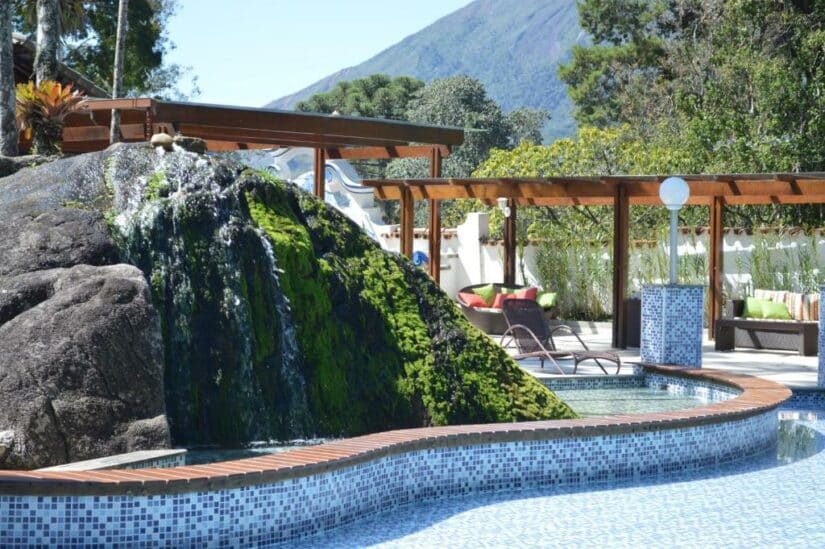 Piscina externa com uma grande rocha coberta de musgo, espreguiçadeiras e uma montanha ao fundo de onde ficar em Teresópolis