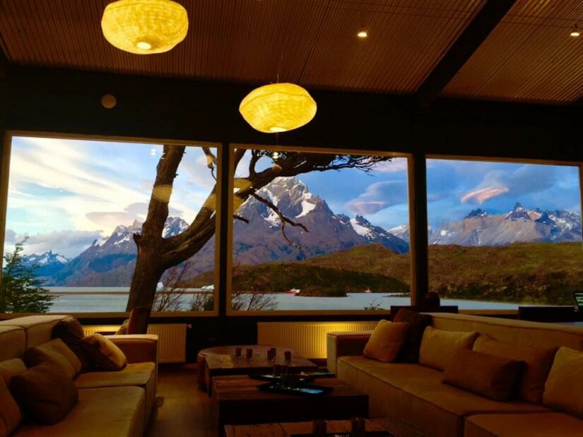 Sala de estar aconchegante com grandes janelas que enquadram montanhas nevadas e um lago ao pôr do sol.