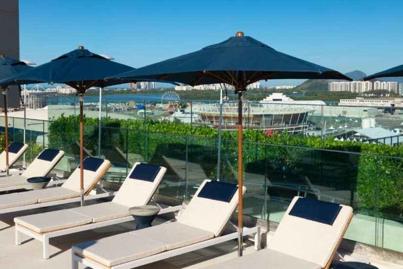 Espreguiçadeiras e guarda-sóis na cobertura da piscina, com uma paisagem urbana e um estádio ao fundo, sob um céu claro.