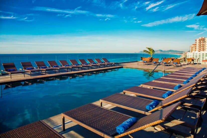 Piscina na cobertura com espreguiçadeiras e toalhas azuis com vista para o oceano e prédios distantes sob um céu claro.